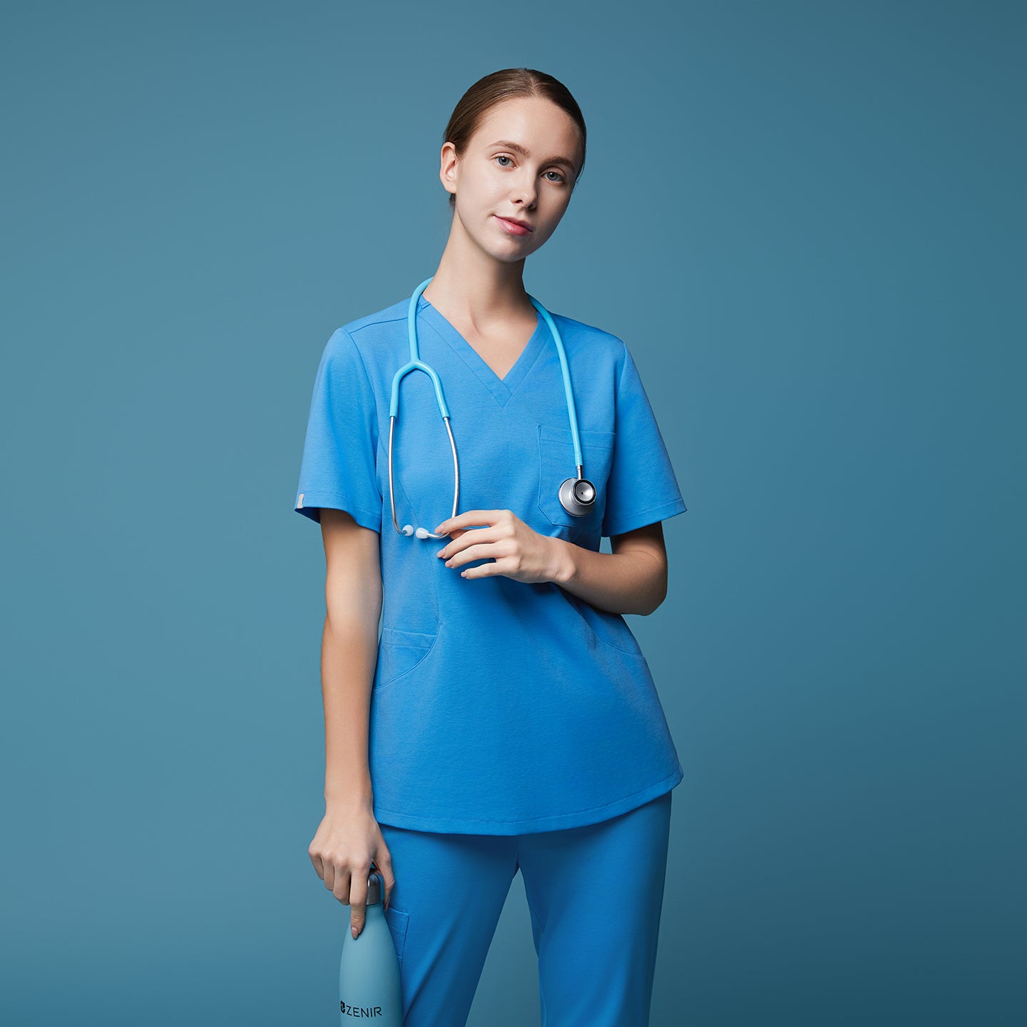 Woman in an aqua blue scrub top and pants, standing confidently with a stethoscope around her neck and holding a matching aqua water bottle, set against a solid aqua background,Aqua Blue