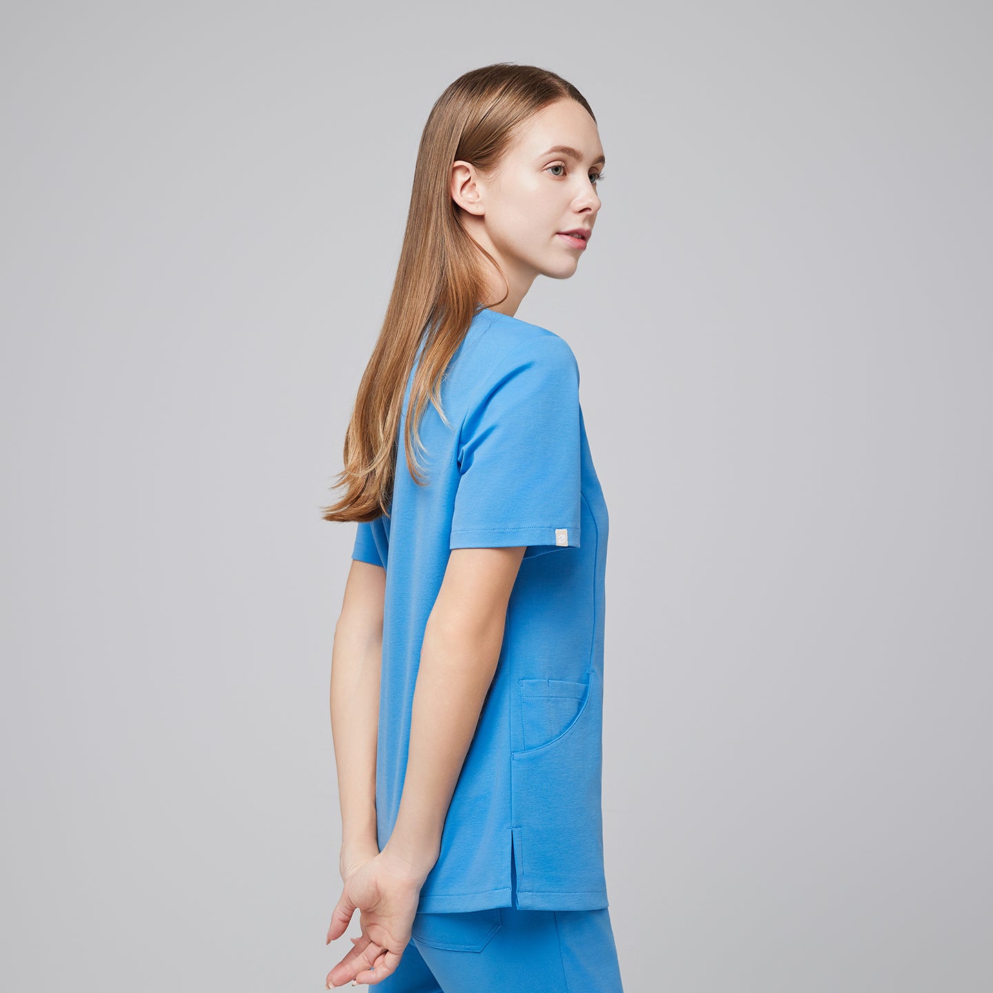 Woman in an aqua blue scrub top and pants, standing in a side pose with hands gently clasped behind her back, looking confidently to the side,Aqua Blue
