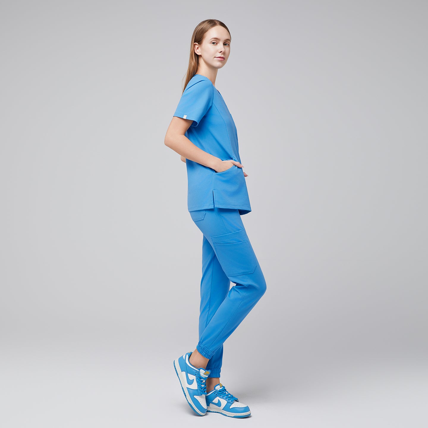Woman wearing an aqua blue scrub top and jogger pants, posing in a side profile with hands in her pockets, paired with matching blue sneakers,Aqua Blue