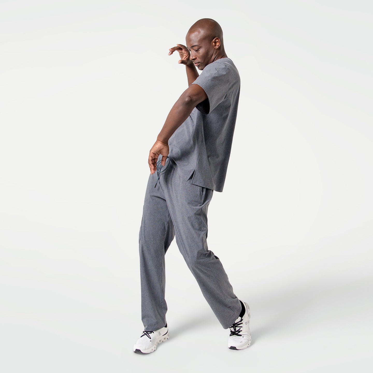 In an Ash Gray scrub set, the male model strikes a dynamic pose with bent arms and a forward lean, showcasing both movement and style.