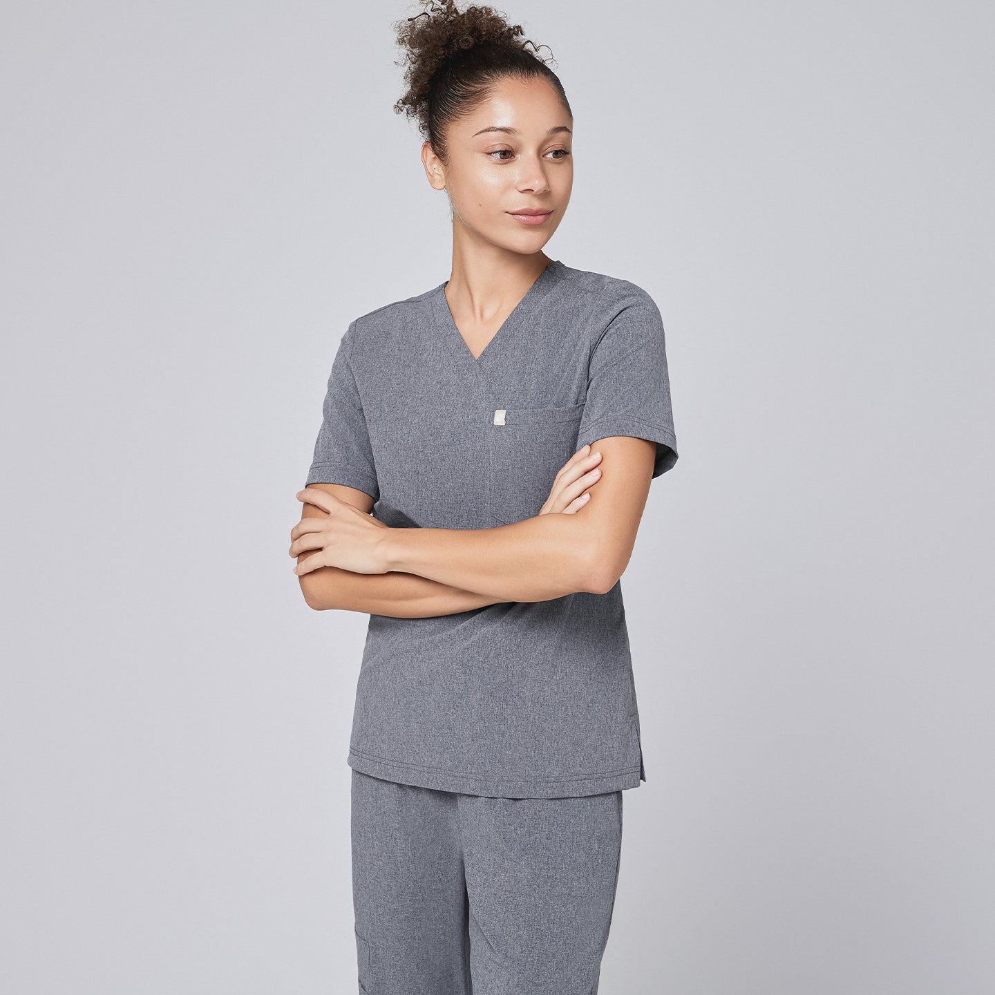 Image of Female healthcare professional in an ash gray scrub set, standing in a rear-side pose with hands in pockets and wearing black sneakers,Ash Gray