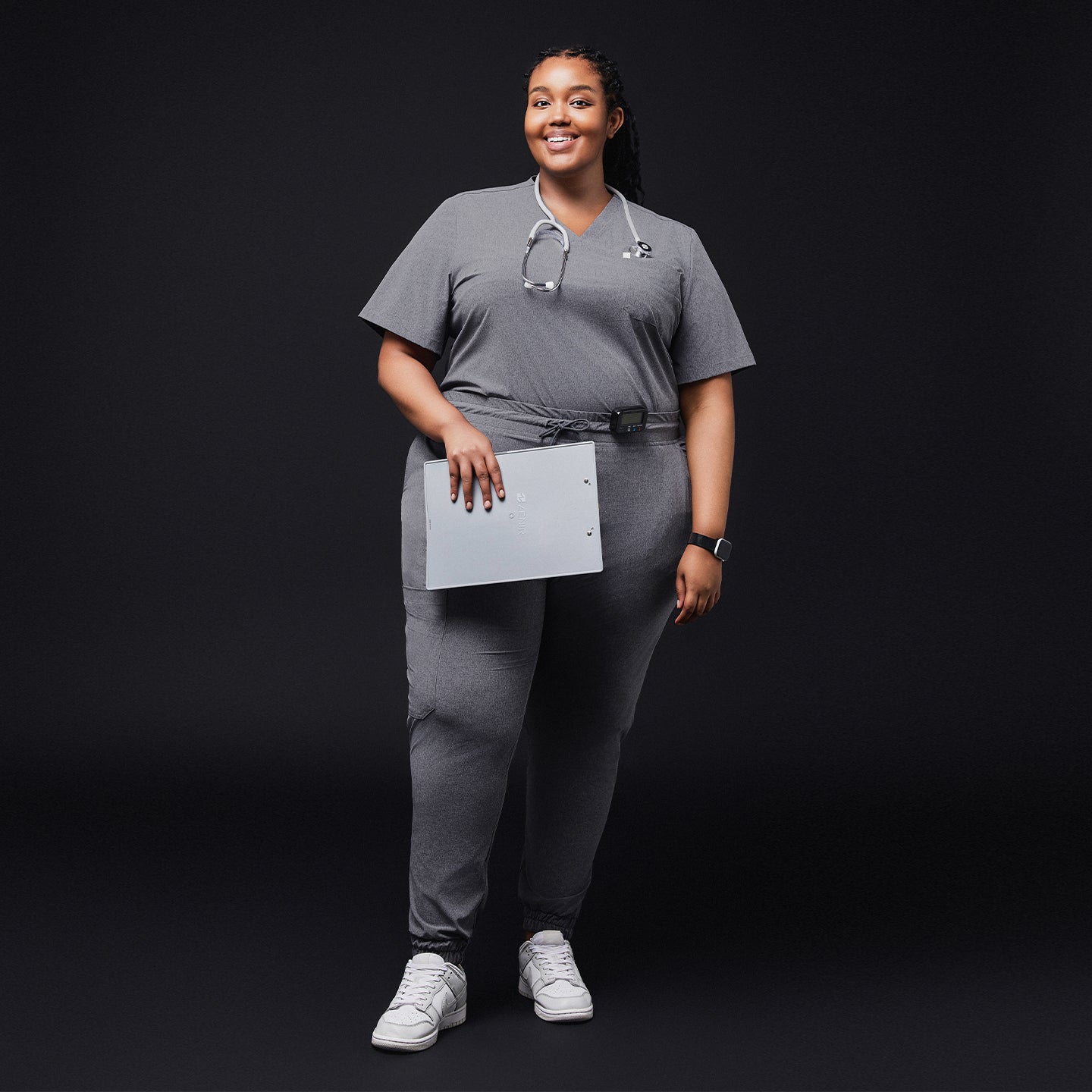 Image of Smiling woman in ash gray scrub set with jogger pants, stethoscope, and white sneakers, holding a clipboard on a black background,Ash Gray