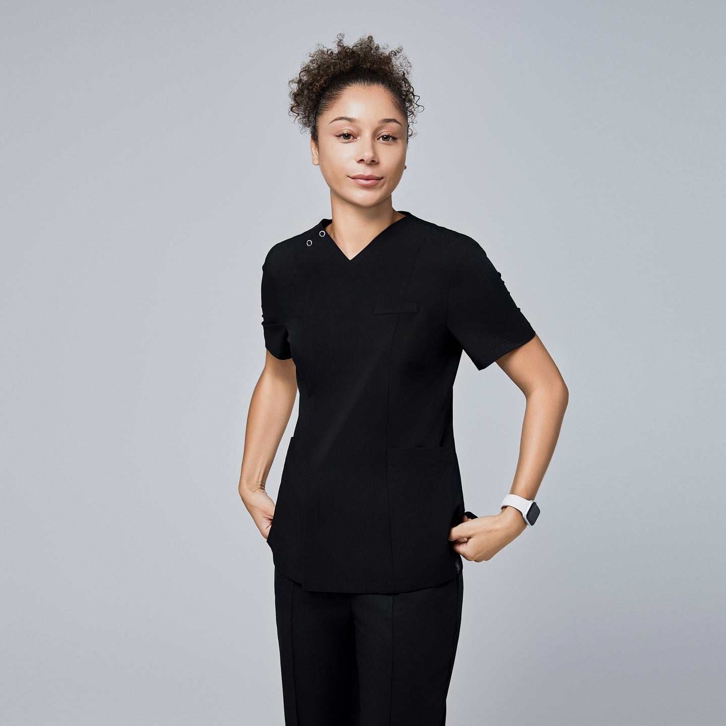 Image of Female model wearing a black V-neck scrub top with a front zipper, short sleeves, and multiple pockets, standing with hands in pockets,Eco Black