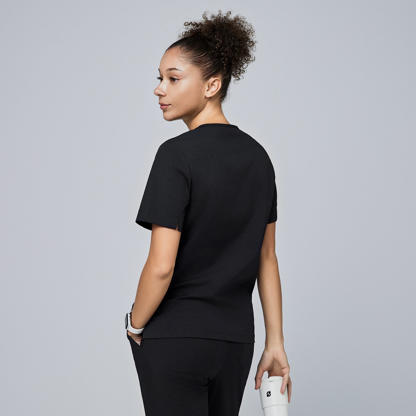 Image of Female model wearing a black scrub top, shown from the back with short sleeves, holding a coffee cup and looking over her shoulder,Eco Black