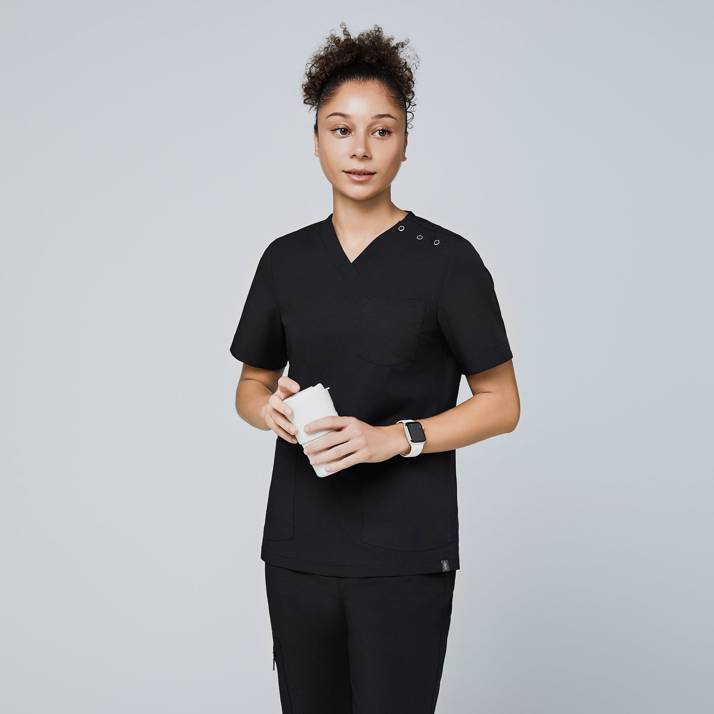 Image of Female model wearing a black V-neck scrub top with two shoulder buttons and short sleeves, holding a coffee cup, and paired with matching black scrub pants,Eco Black