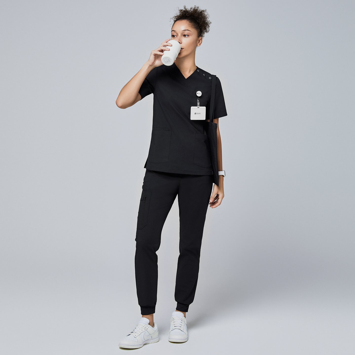Image of Female model wearing a black V-neck scrub top with shoulder buttons, black jogger scrub pants, a name badge, and a tote bag, drinking from a reusable cup,Eco Black