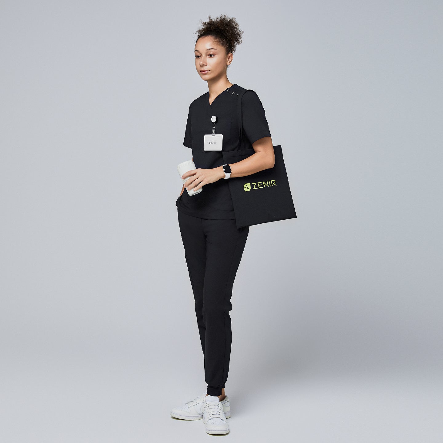 Image of Female model wearing a black V-neck scrub top with shoulder buttons, black scrub pants, holding a coffee cup and wearing a name badge, with a black Zenir tote bag over her shoulder,Eco Black