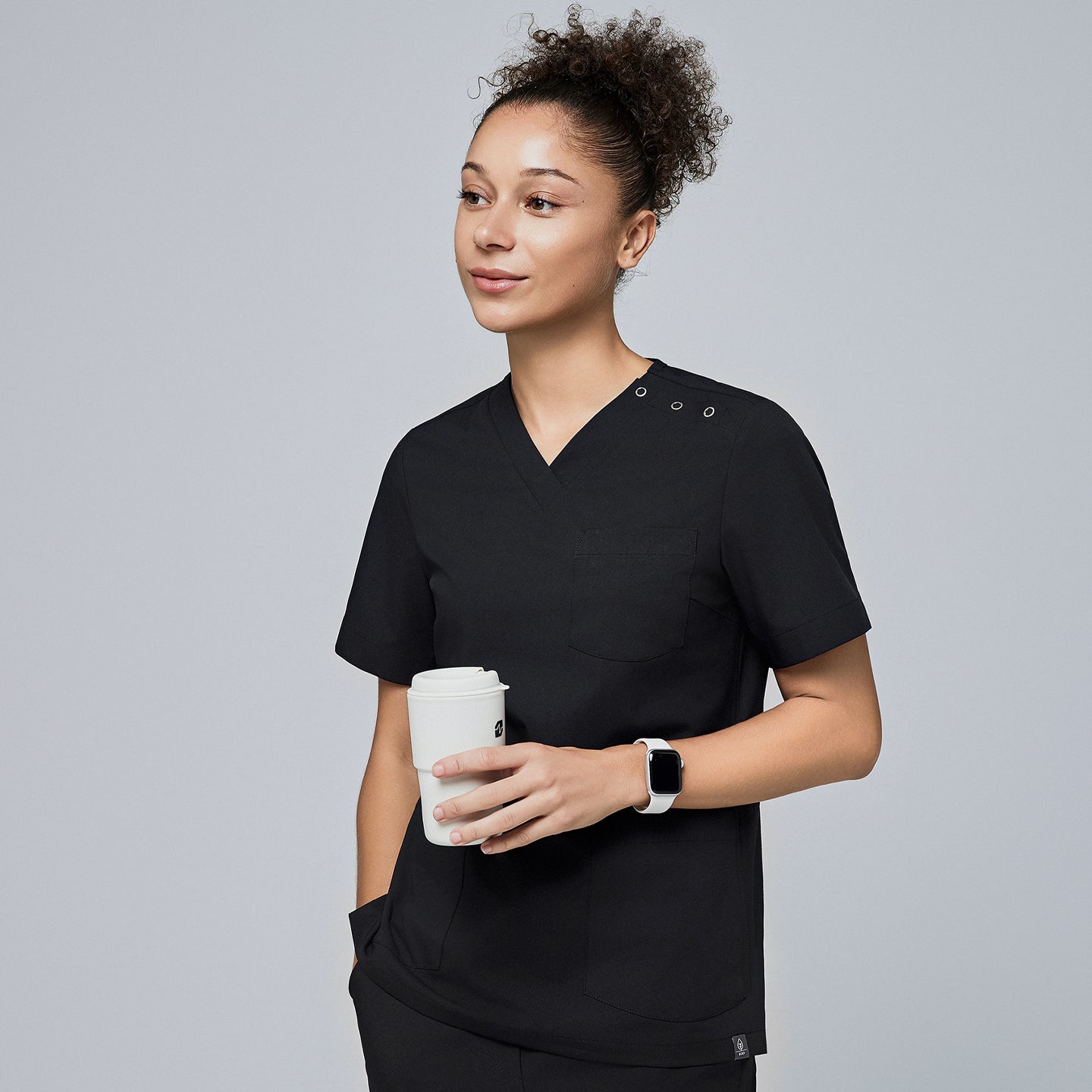 Female model wearing a black V-neck scrub top with two shoulder buttons and short sleeves, holding a coffee cup and looking to the side,Eco Black