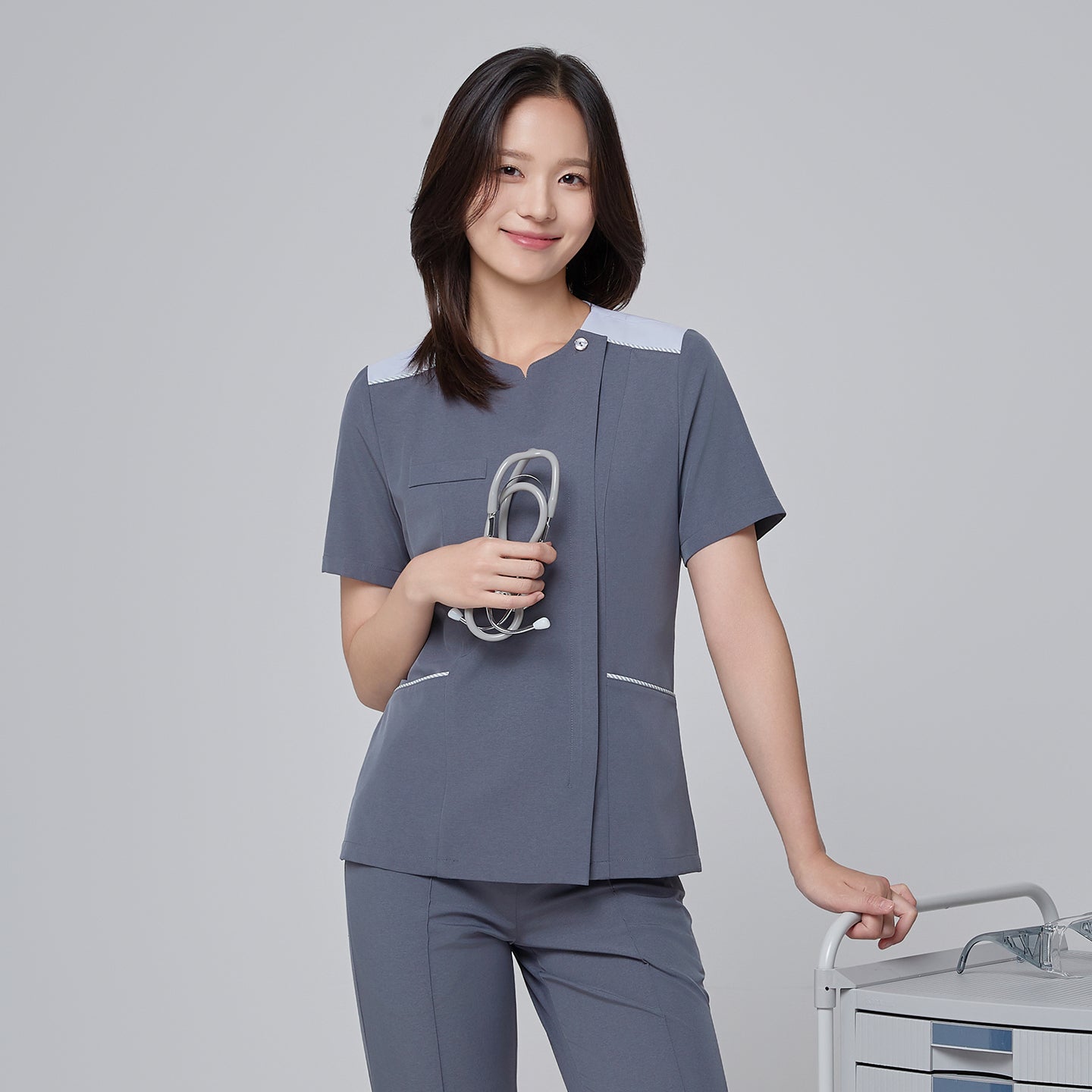Lifestyle image of a healthcare professional wearing the TUL OS-202 Women's One Button Front Zipper Scrub Top in Blue Gray in a clinical setting. The model is shown interacting with equipment in a softly blurred hospital background, emphasizing the top’s modern design and professional fit.