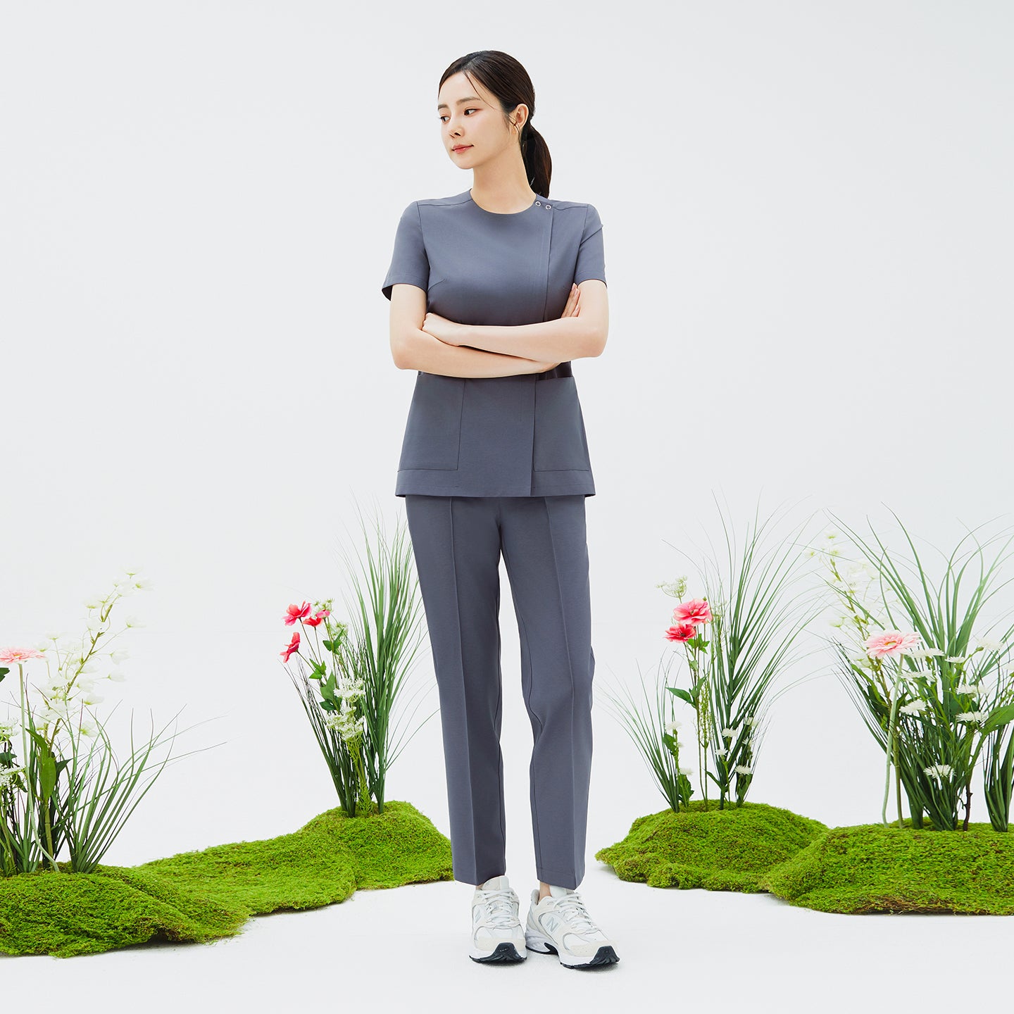Image of Model wearing TUL OS-212 Blue Gray scrub top with arms crossed, set against a nature-inspired backdrop, emphasizing a clean, tailored silhouette.