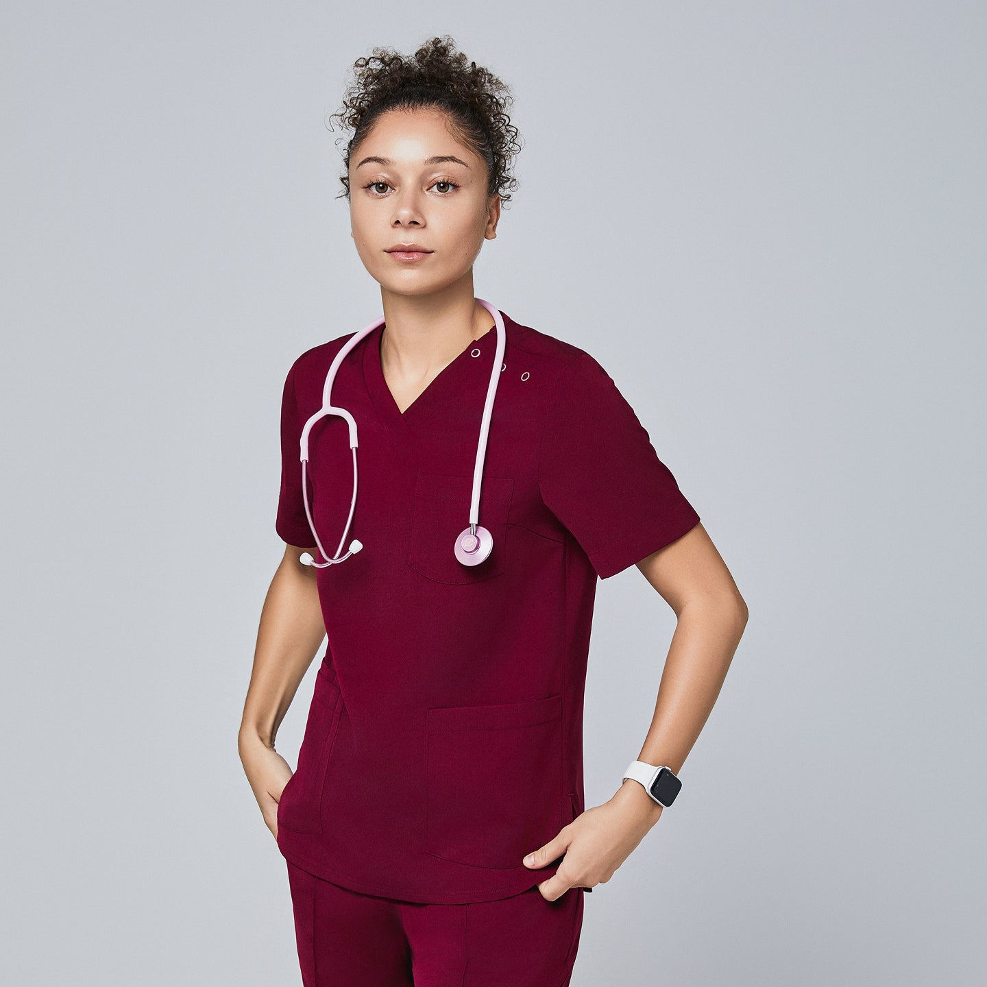 Image of Female model wearing a burgundy V-neck scrub top with a three-button detail and short sleeves, accessorized with a pink stethoscope and paired with matching scrub pants,Burgundy