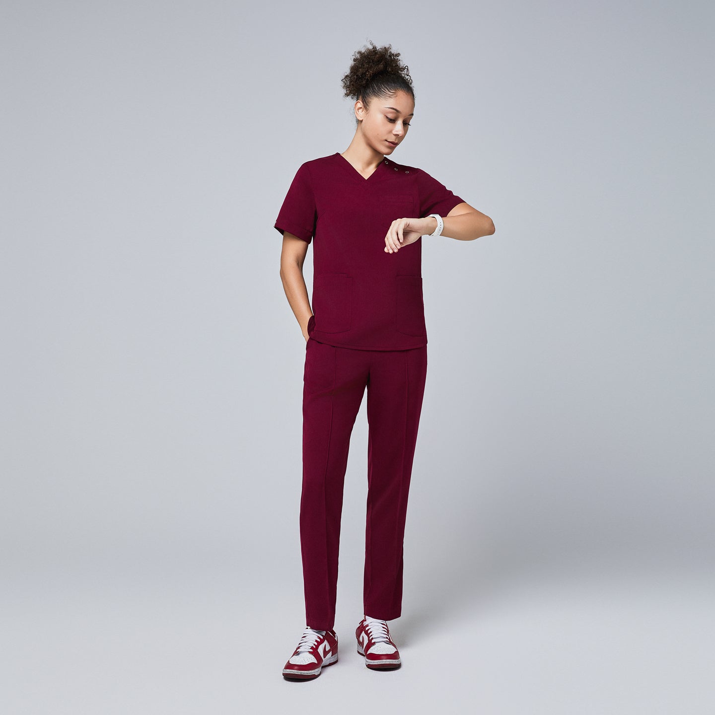 Image of Female model wearing a burgundy V-neck scrub top with a three-button detail, short sleeves, and matching burgundy scrub pants, checking the time on her smartwatch,Burgundy