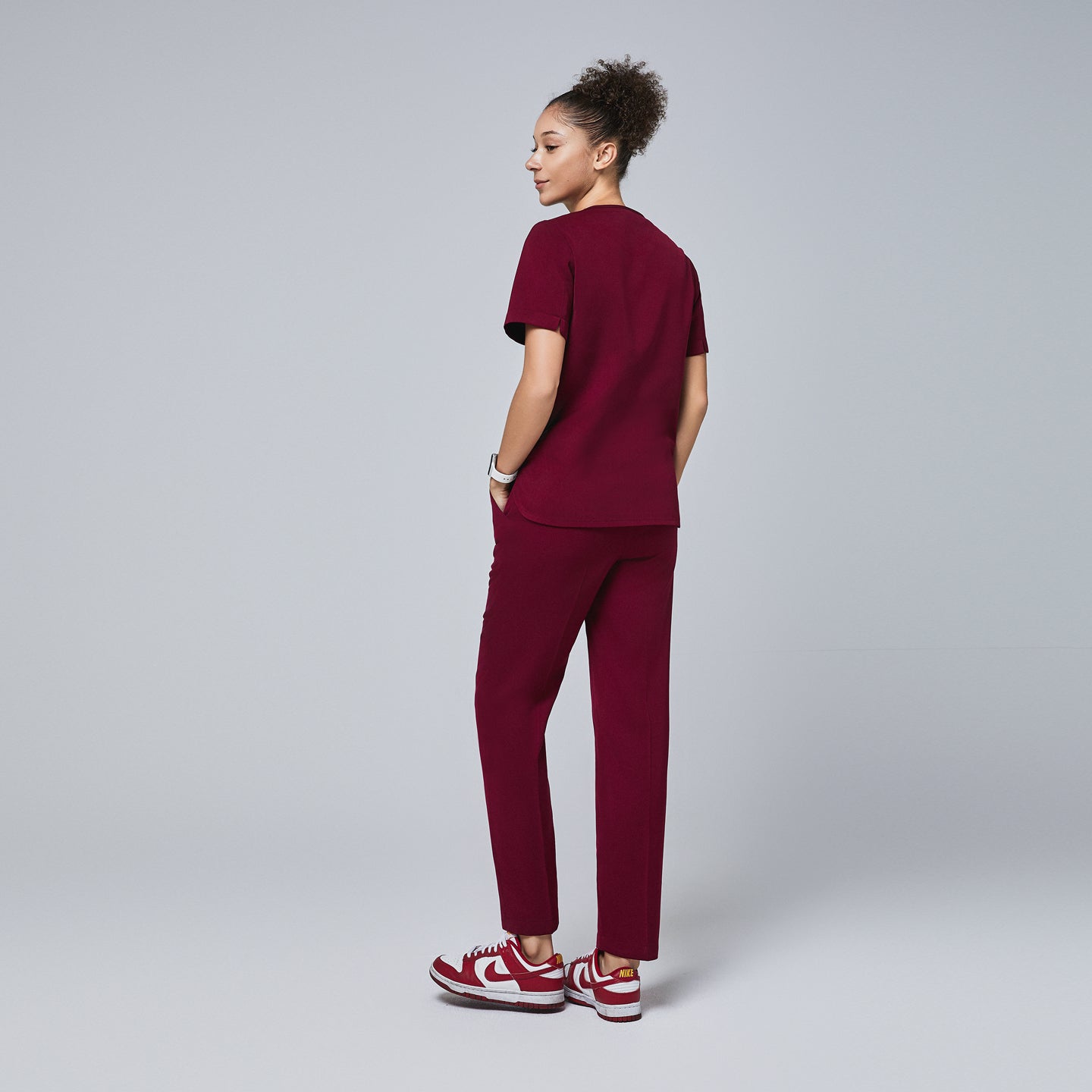 Image of Female model wearing a burgundy scrub top with short sleeves and matching pants, shown from the back with hands in pockets, paired with burgundy and white sneakers,Burgundy