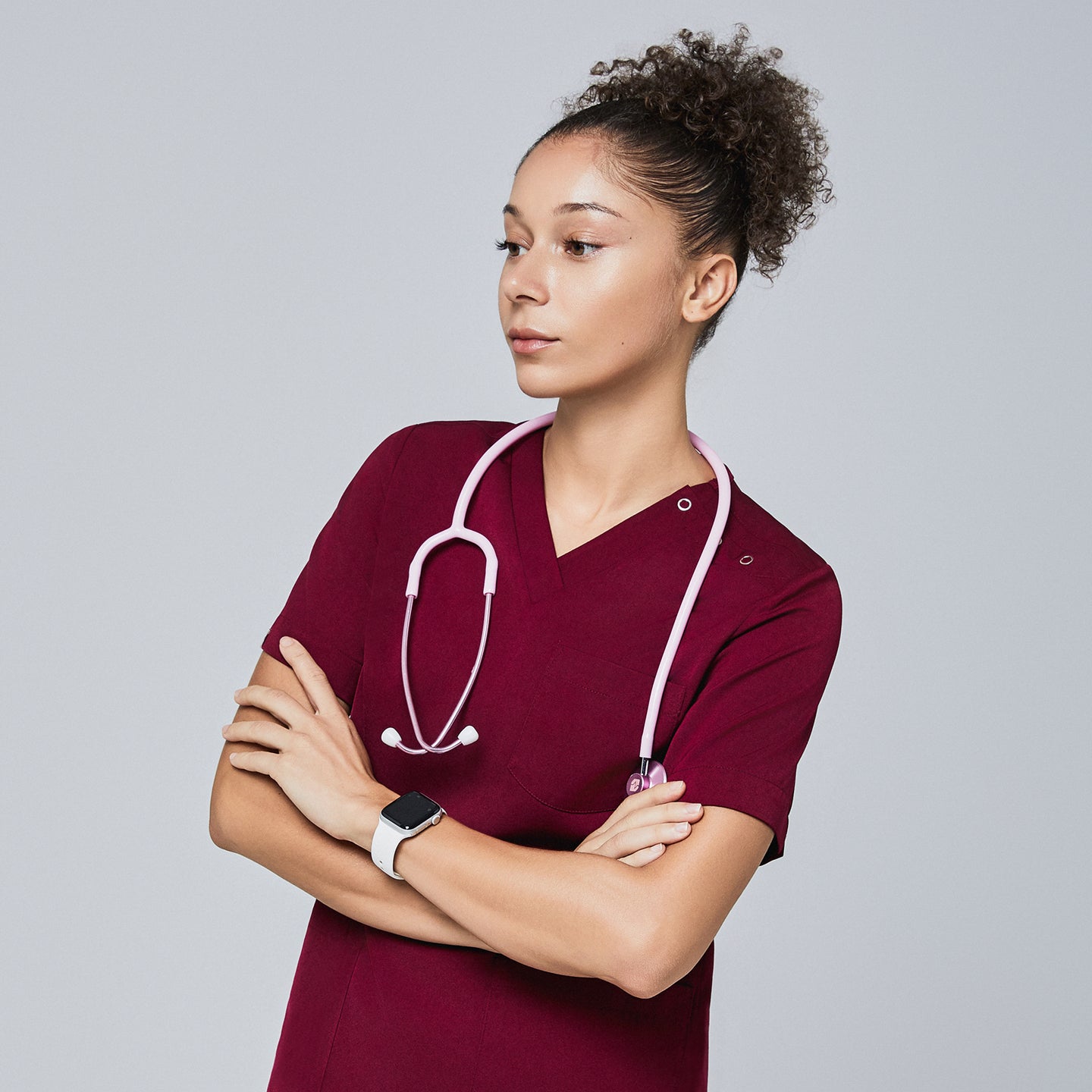 Image of Female model wearing a burgundy V-neck scrub top with three-button detail, accessorized with a pink stethoscope around her neck and a smartwatch, standing with arms crossed and looking to the side, Burgundy