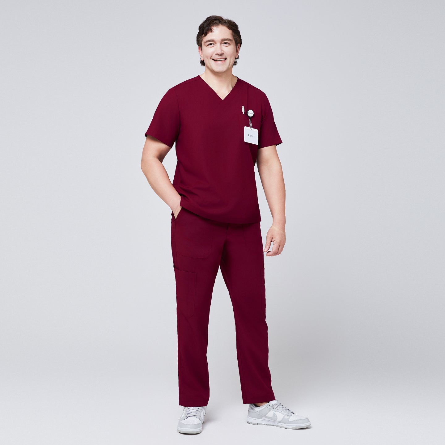Image of Man wearing a burgundy V-neck scrub top with badge and pocket accessories, paired with matching burgundy scrub pants and white sneakers,Burgundy
