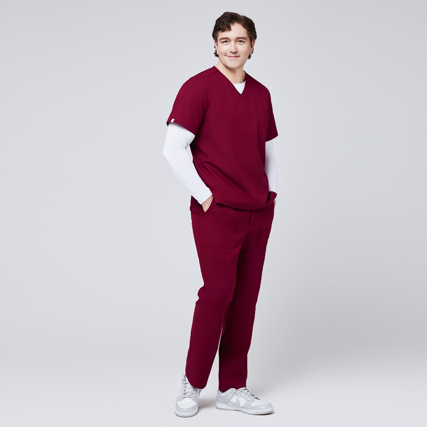 Man wearing a burgundy V-neck scrub top layered over a white long-sleeve undershirt, paired with matching scrub pants and white sneakers,Burgundy