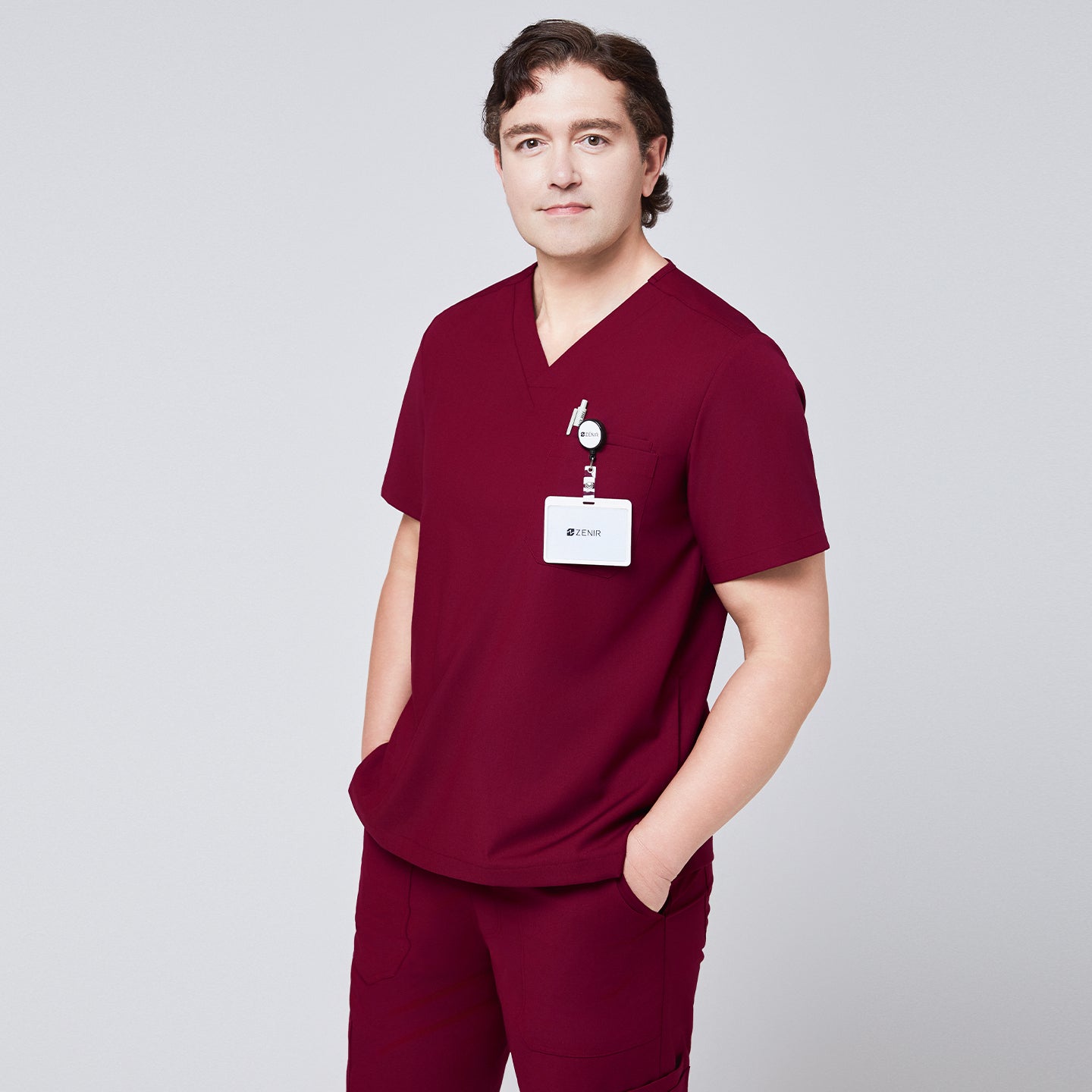 Man in a burgundy V-neck scrub top with badge holder and front pocket, paired with matching scrub pants, hands in pockets,Burgundy
