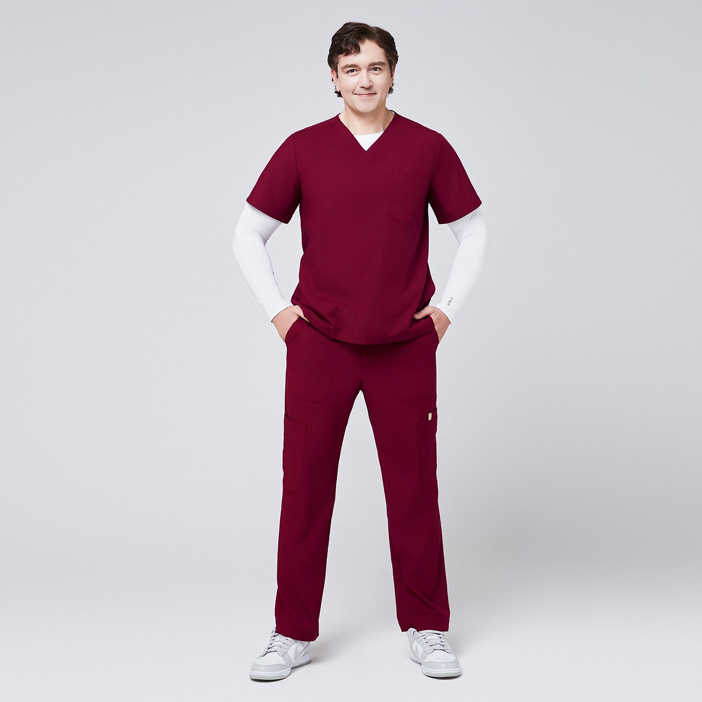 Image of Man wearing a burgundy V-neck scrub top layered over a white long-sleeve undershirt, paired with matching scrub pants and white sneakers,Burgundy