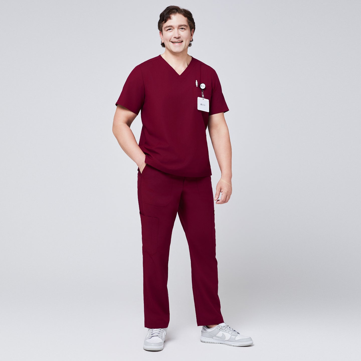 Image of Man wearing burgundy V-neck scrub top and 8-pocket straight scrub pants, paired with white sneakers and a name badge,Burgundy