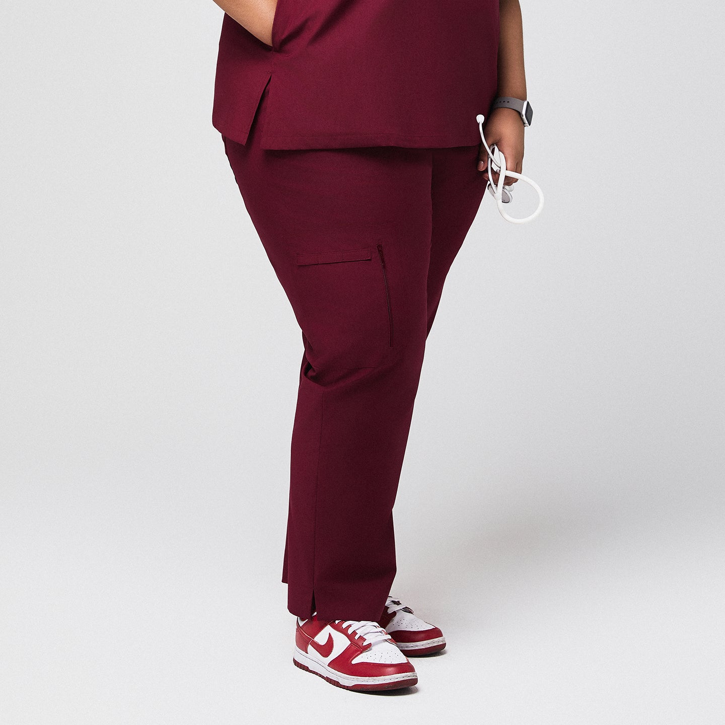 Image of Side view of burgundy scrub pants with zipper pocket and split hem, paired with red sneakers,Burgundy