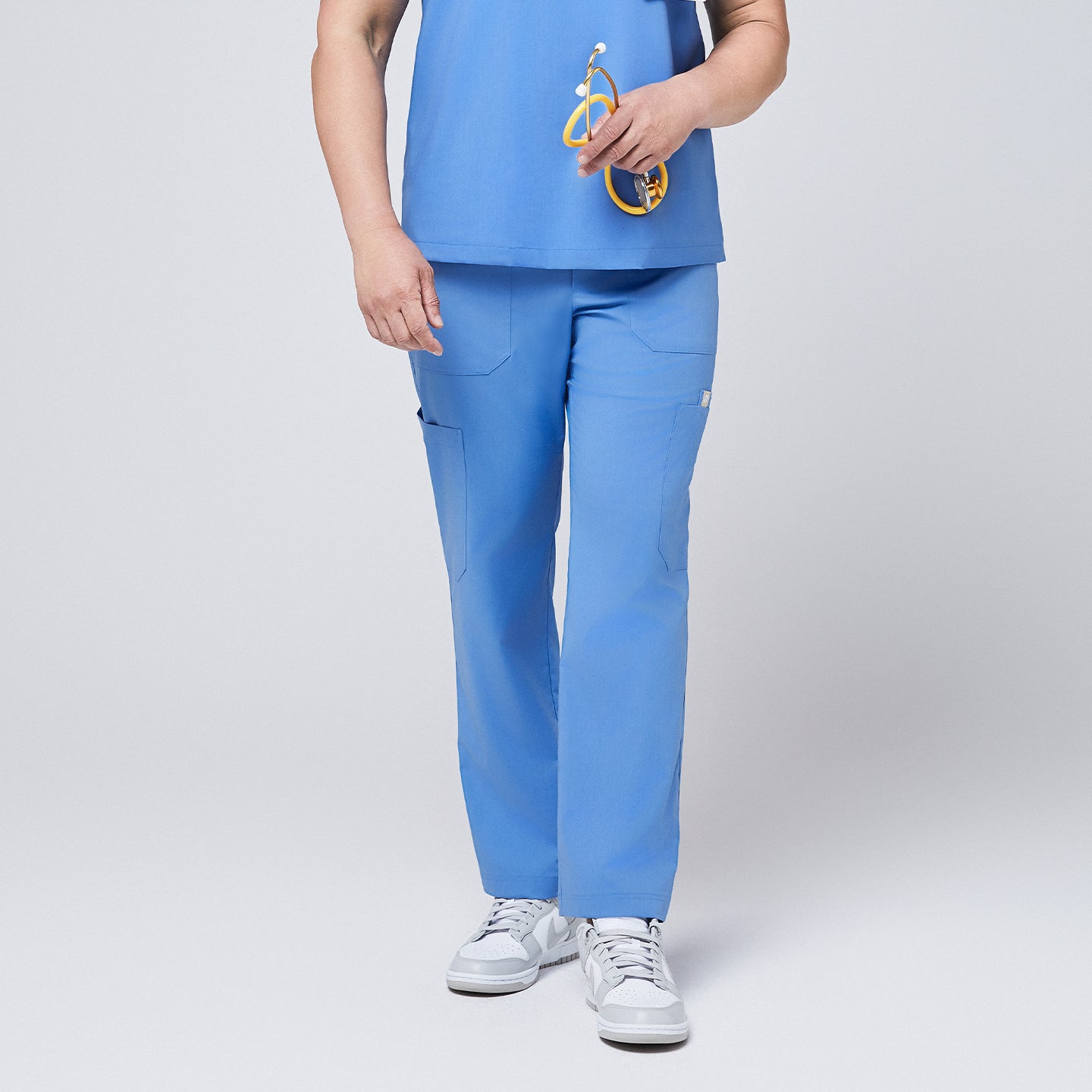 Image of Close-up view of the lower half of a person wearing ceil blue straight scrub pants with multiple utility pockets, paired with white sneakers, holding a yellow stethoscope,Ceil Blue