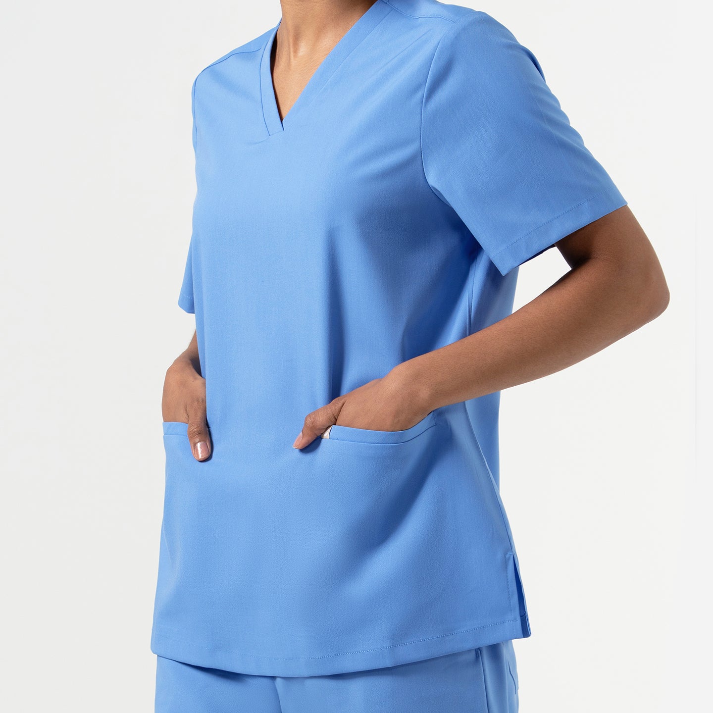 Woman wearing a Ceil Blue double-pocket scrub top, shown from the side with hands in the front pockets, highlighting the smooth V-neck design, short sleeves, and soft flexible fabric.