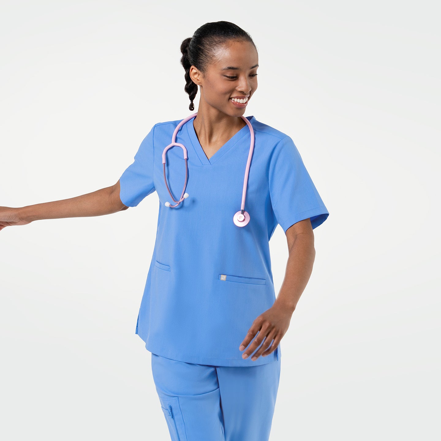 Image of Smiling in the Women's Ceil Blue Double Pocket Scrub Top and pants, pink stethoscope around the neck, mid-motion, full body view