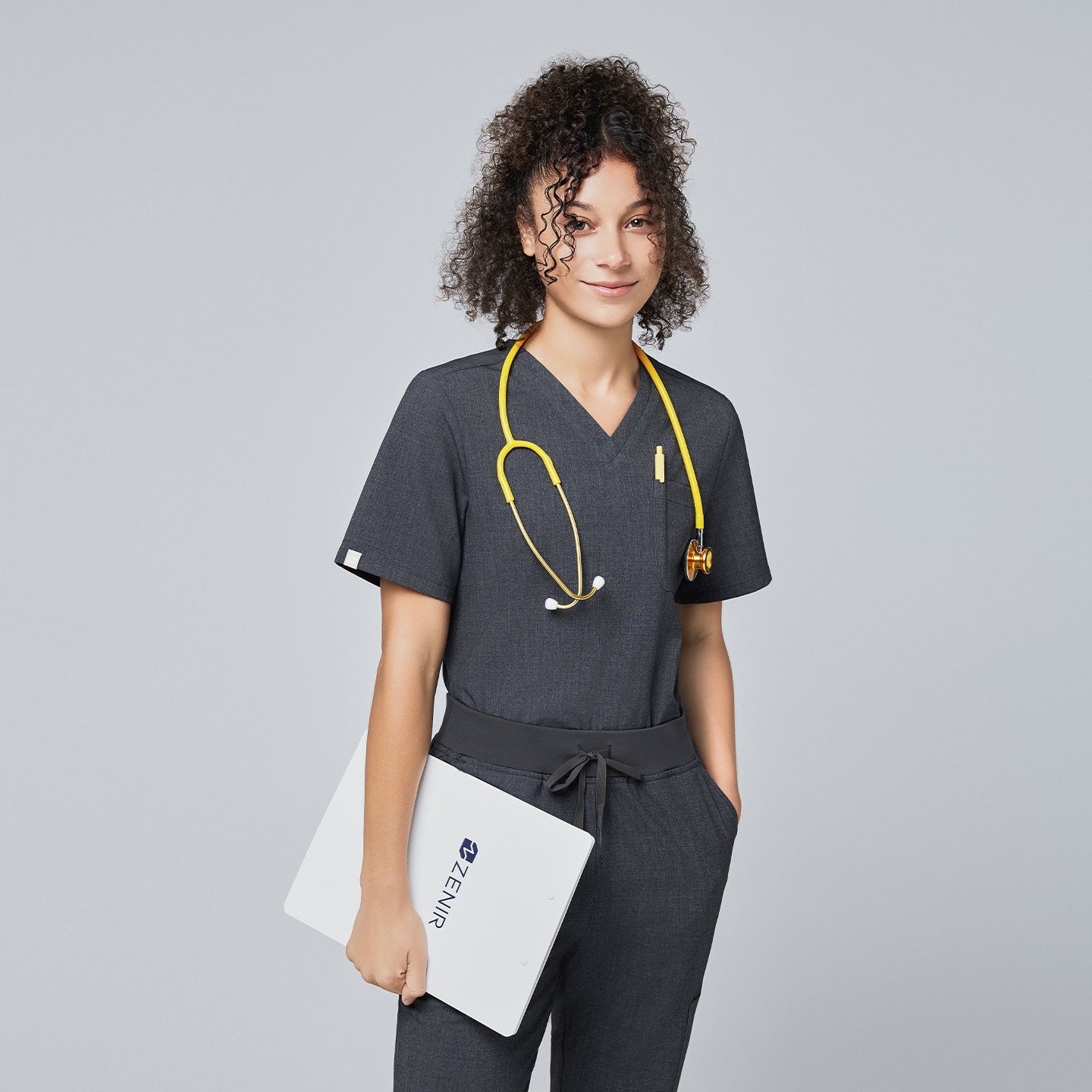 Image of Model wearing Zenir charcoal gray 3-pocket scrub top with a yellow stethoscope and holding a clipboard,Charcoal Gray
