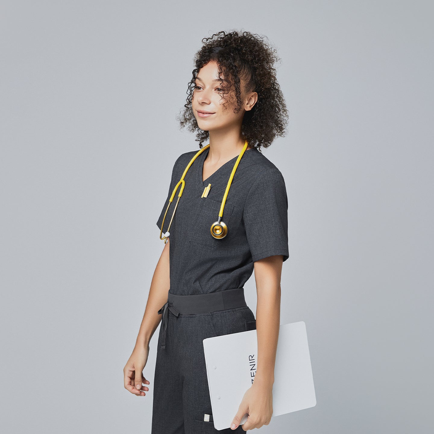 Image of Side view of a model wearing Zenir charcoal gray 3-pocket scrub top with a yellow stethoscope and holding a clipboard,Charcoal Gray