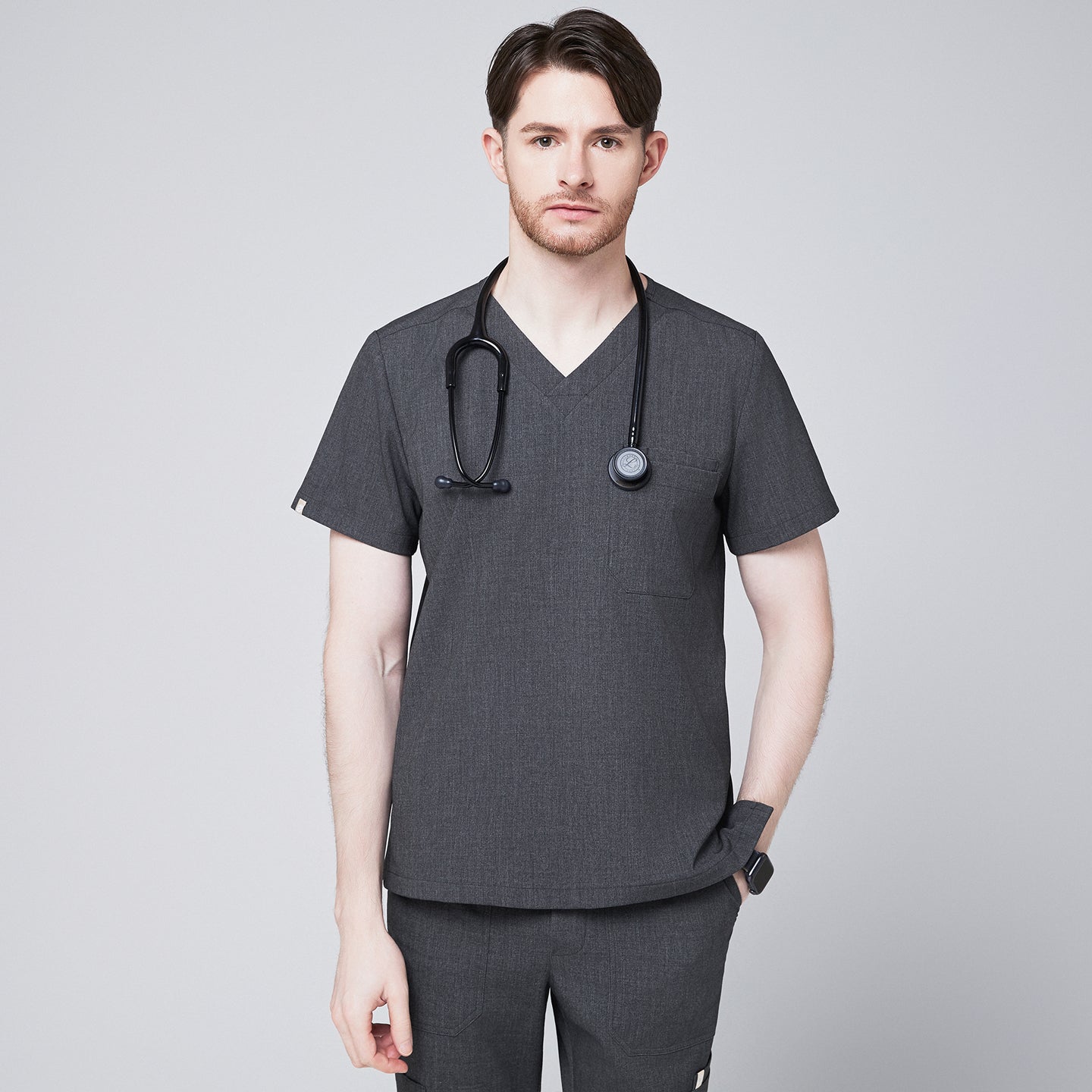 Image of Male healthcare professional wearing a charcoal gray V-neck scrub top with three pockets and black stethoscope around neck, standing against a plain background,Charcoal Gray