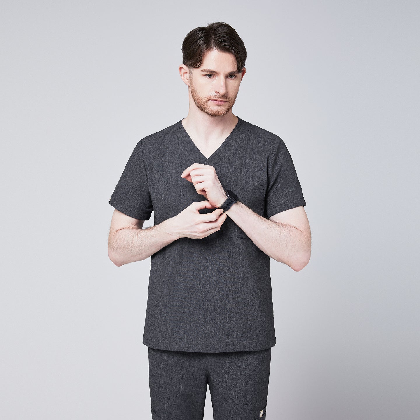 Image of Male healthcare professional wearing charcoal gray V-neck scrub top and pants, adjusting his smartwatch, standing against a plain background,Charcoal-Gray