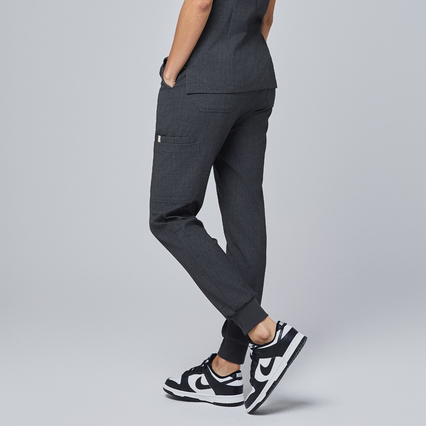 Image of Side view of charcoal gray jogger scrub pants featuring a side pocket, elastic waistband with a drawstring, and cuffed ankles, paired with white sneakers,Charcoal Gray