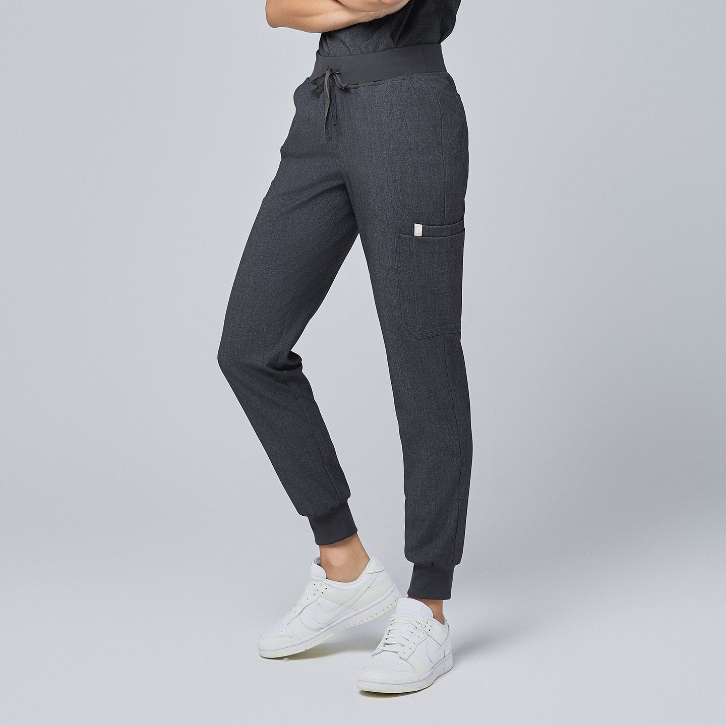Image of Side view of charcoal gray jogger scrub pants featuring a side pocket, elastic waistband with a drawstring, and cuffed ankles, paired with white sneakers,Charcoal Gray