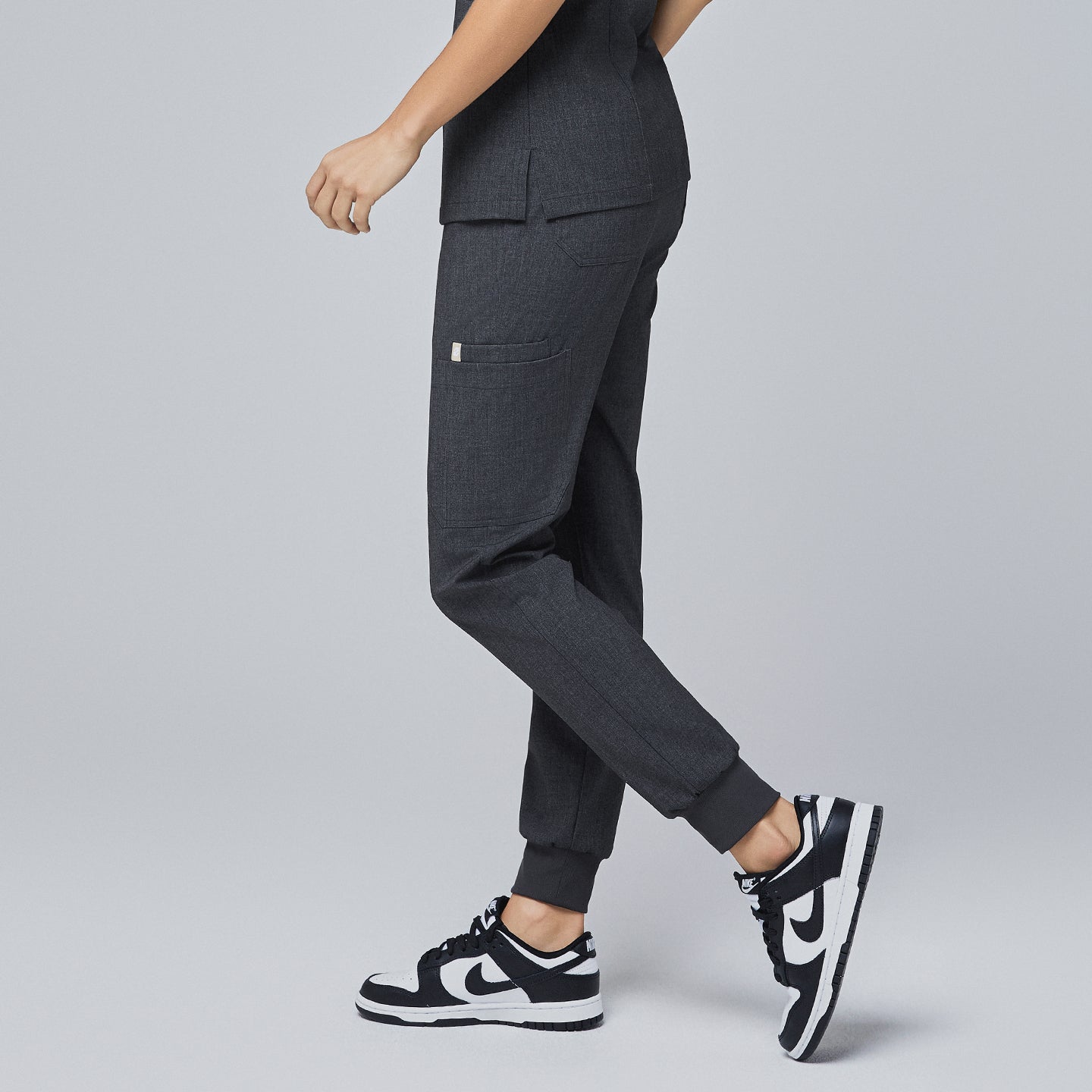 Image of Side angle of charcoal gray jogger scrub pants with a side pocket, elastic cuffs, and a tapered fit, paired with black-and-white sneakers,Charcoal Gray