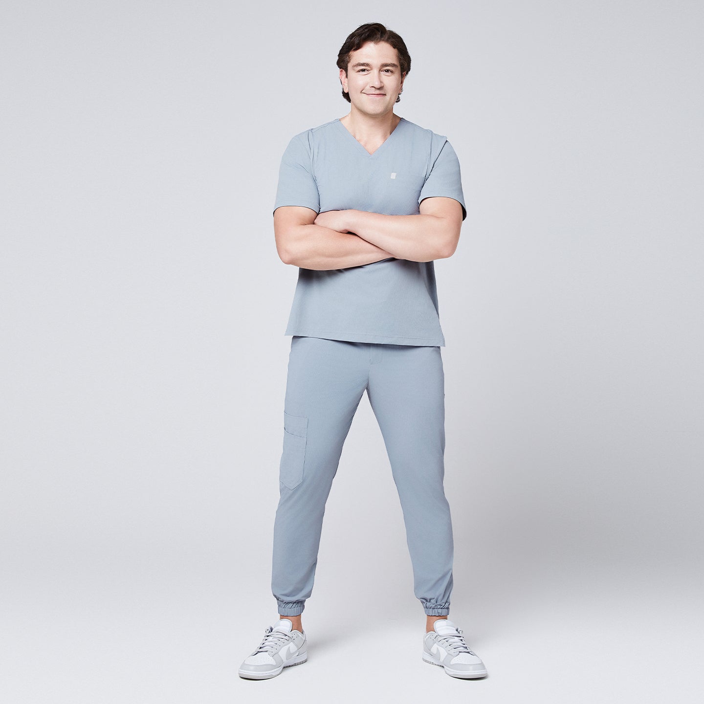 Image of Full-body view of a man in a cool gray scrub set with jogger pants, arms crossed, and wearing white sneakers,Cool Gray