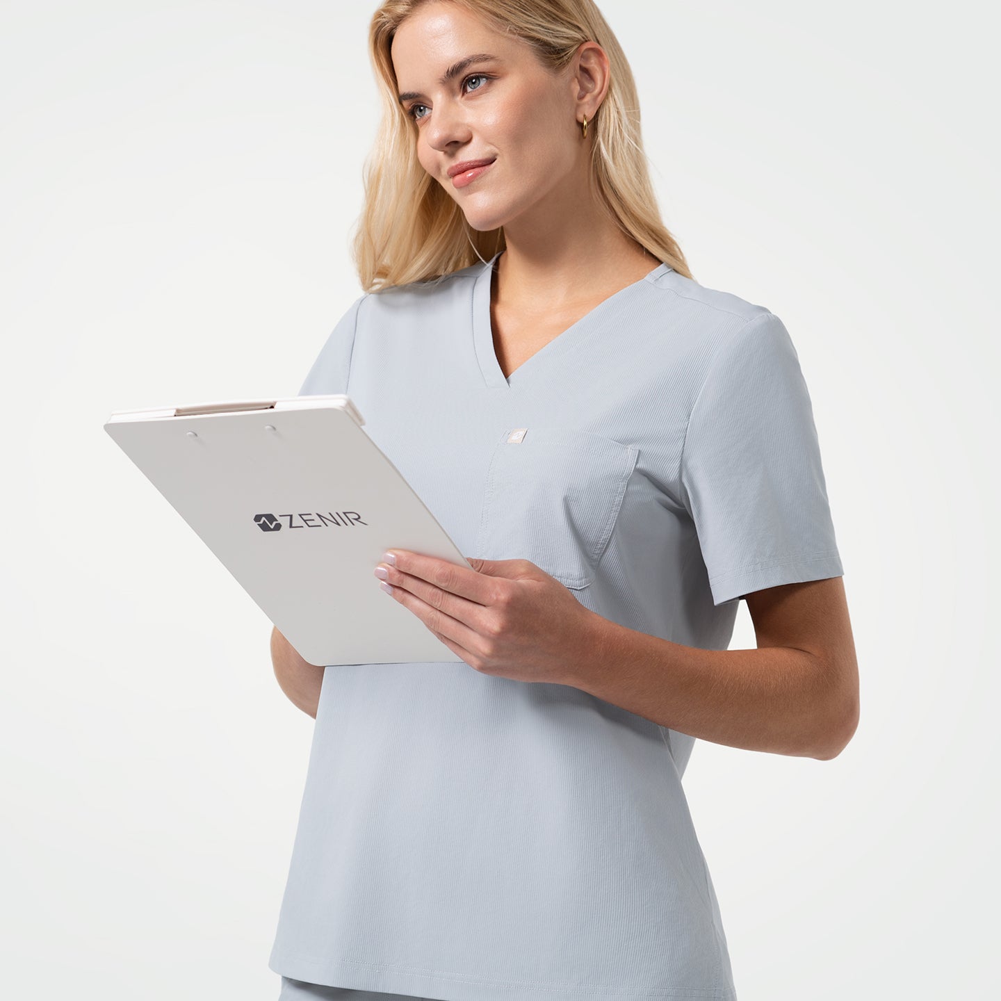 Holding a white clipboard marked with the Zenir logo, the model showcases a Cool Gray V-neck scrub top with short sleeves and chest pocket, styled with matching scrub pants against a clean light background.