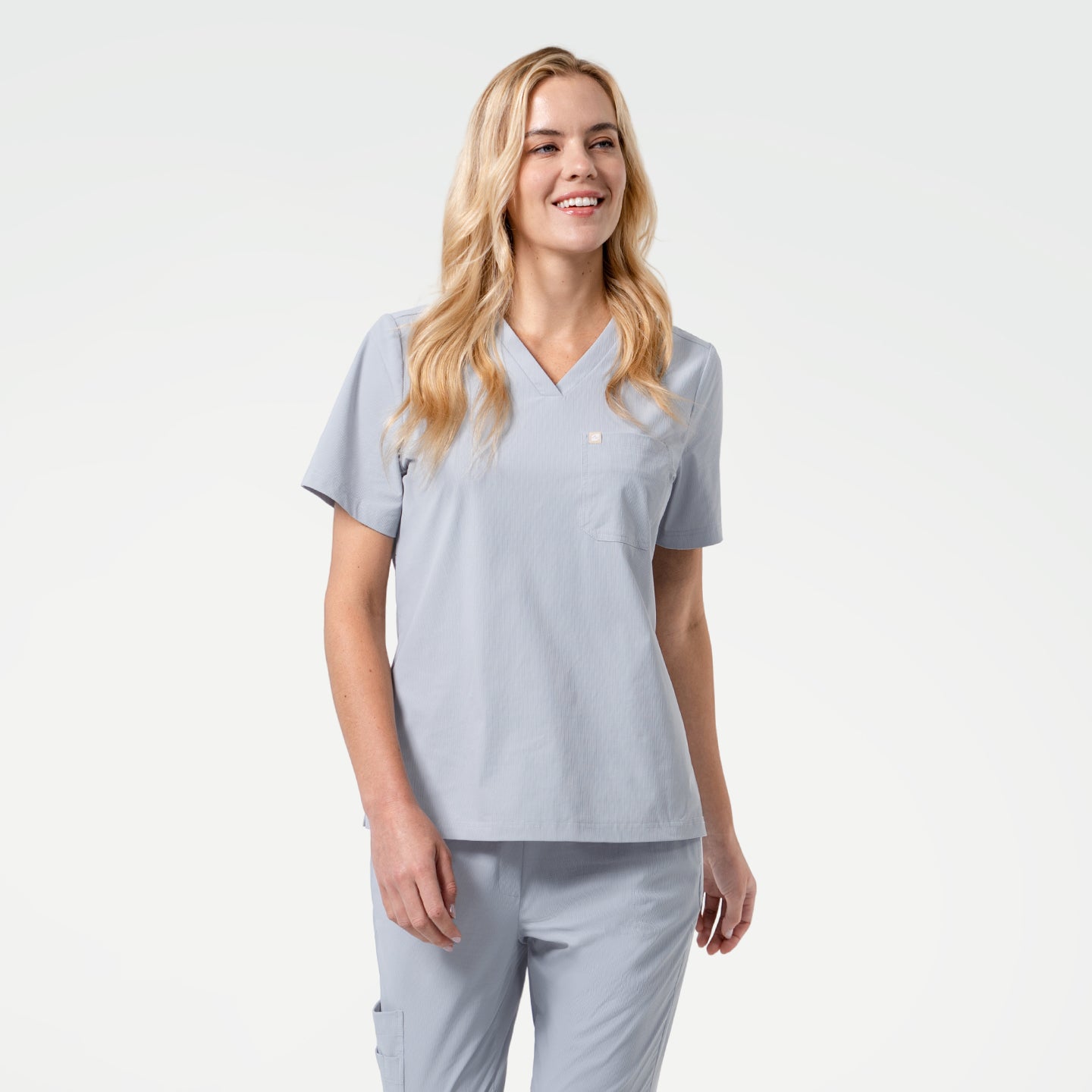 The model wears a Cool Gray single-pocket scrub top featuring a clean V-neckline and short sleeves for a neat, professional look. The lightweight gray fabric falls smoothly and offers easy movement throughout the shift. A single chest pocket provides simple, functional storage, while the matching Cool Gray scrub pants complete the coordinated, minimal, and modern uniform style.