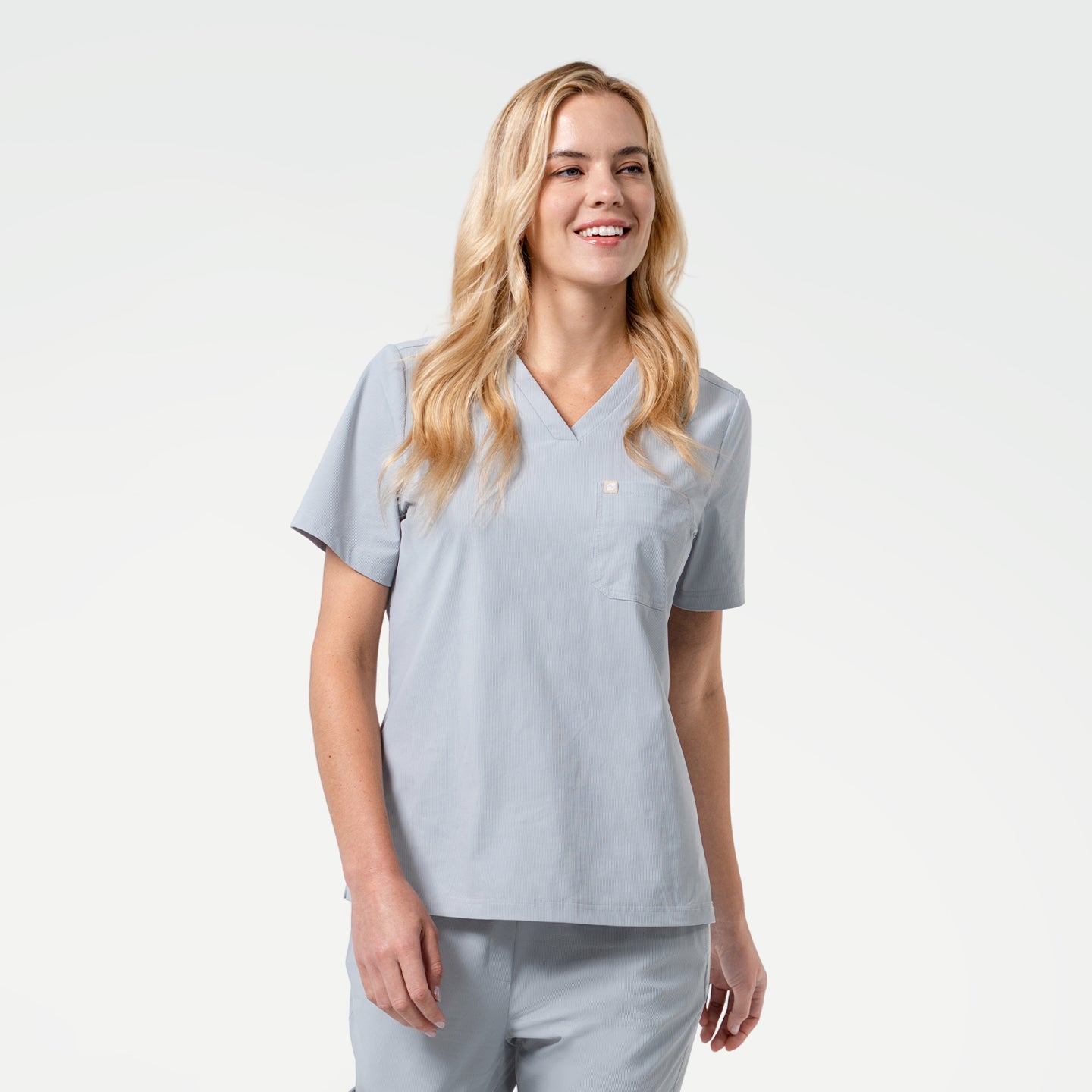 With a bright smile, the model wears a Cool Gray V-neck scrub top featuring short sleeves and a chest pocket, paired with matching scrub pants against a clean light background.