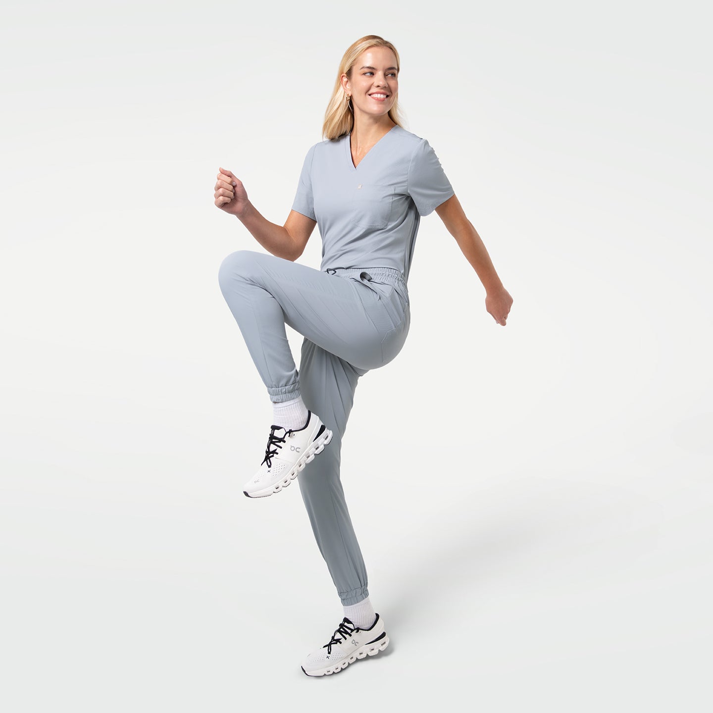 Image of Model wearing a Cool Gray single-pocket scrub top and matching jogger scrub pants, lifting one knee in a dynamic pose to show stretch and mobility. The lightweight fabric moves easily, and the outfit maintains a sleek, modern medical-uniform look.
