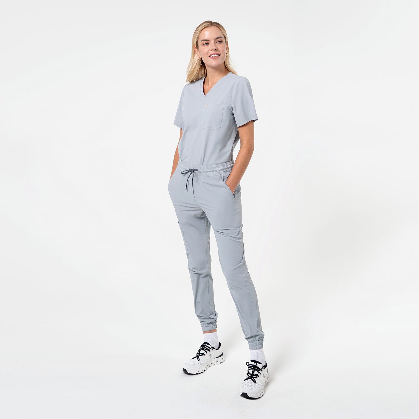 Model wearing a Cool Gray single-pocket scrub top paired with matching jogger scrub pants, standing with hands in pockets. The set has a clean, minimal look with a relaxed fit that highlights comfort and everyday mobility.