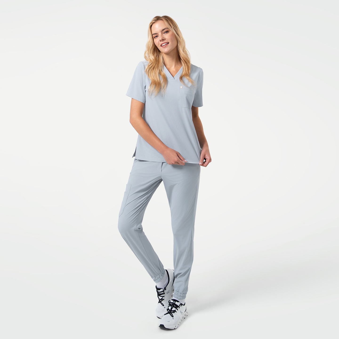 Smiling model stands with one leg crossed in front of the other, wearing a Cool Gray V-neck scrub top with short sleeves and matching jogger-style scrub pants, paired with white sneakers against a clean light background.