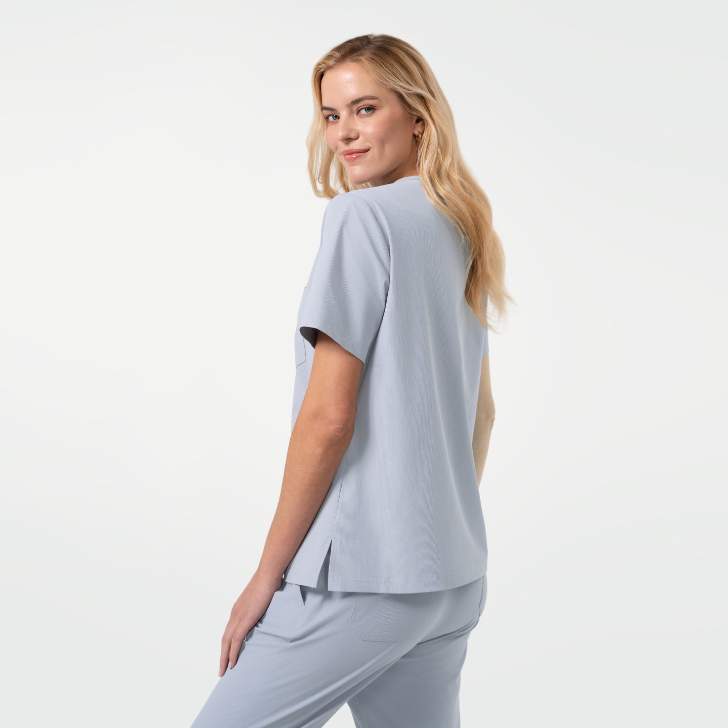 Image of Model turns slightly to the side with a subtle smile, showcasing the back and side view of a Cool Gray V-neck scrub top with short sleeves and side vents, paired with matching scrub pants against a clean light background.