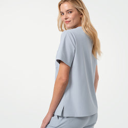 Image of Model turns slightly to the side with a subtle smile, showcasing the back and side view of a Cool Gray V-neck scrub top with short sleeves and side vents, paired with matching scrub pants against a clean light background.