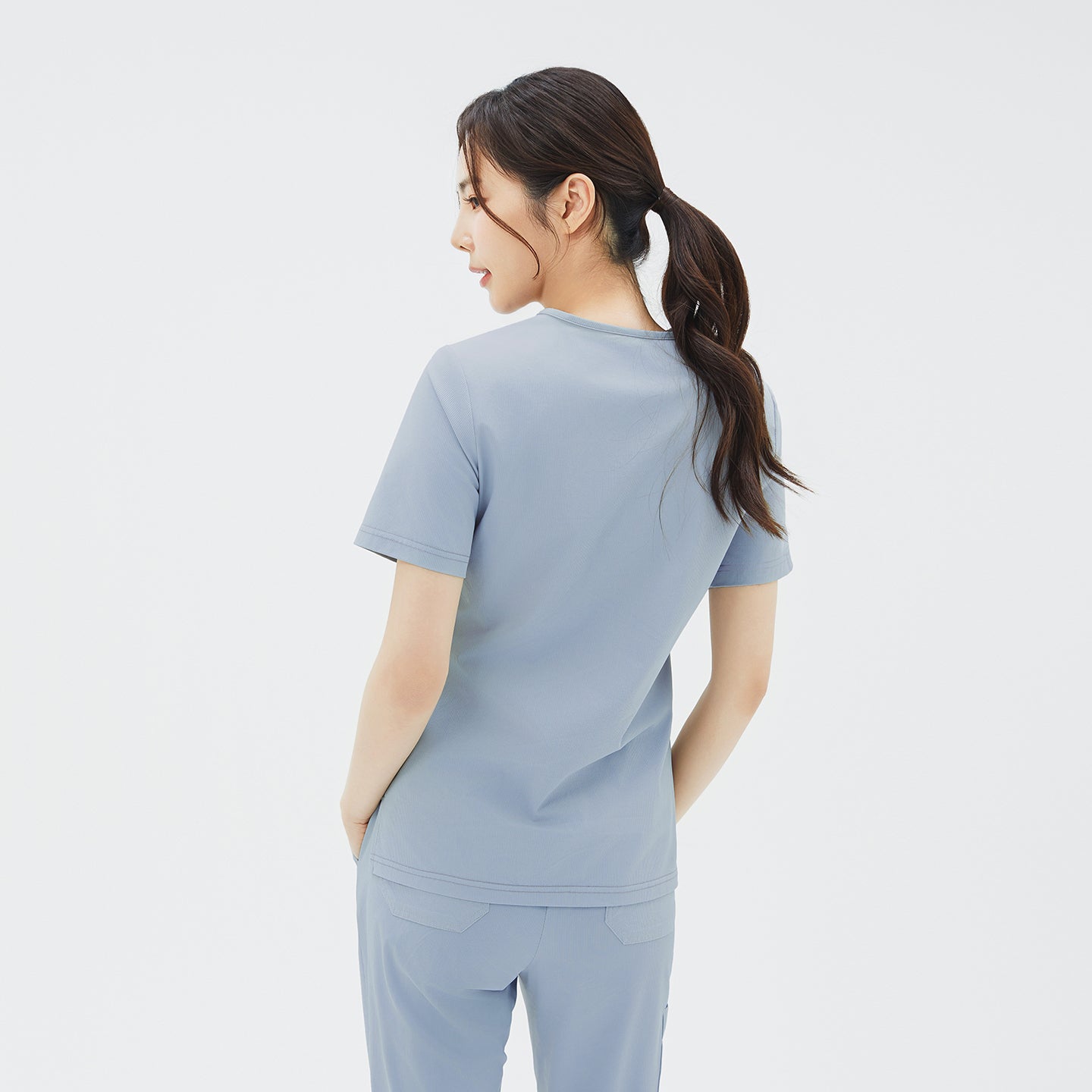 Image of A back three‑quarter shot of the model with her hair in a ponytail and hands in her scrub pants’ back pockets, emphasizing the clean lines and soft drape of the Cool Gray scrub top across the shoulders.