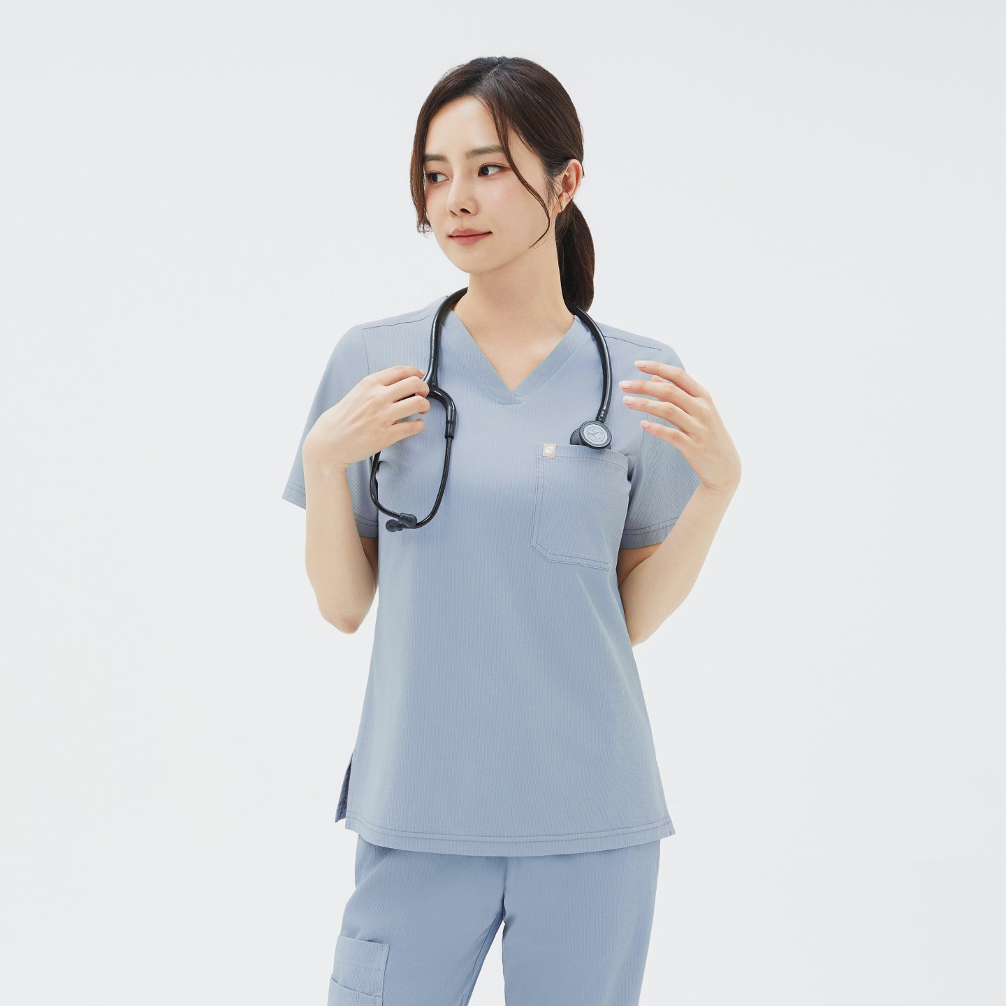 A three-quarter front view of a female healthcare professional wearing the Women’s Single Pocket Scrub Top in Cool Gray with matching scrub pants and a black stethoscope around her neck, reaching up to adjust the tubing while looking off to her left.