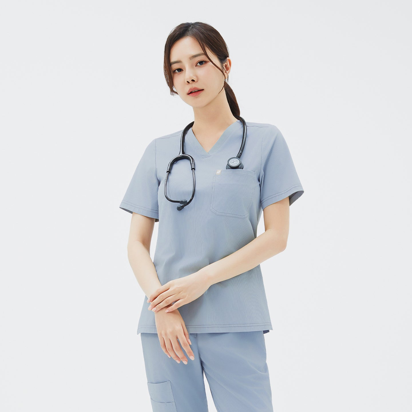 A straight‑on shot of the model standing in the Cool Gray scrub top and pants, hands loosely clasped at her waist and the stethoscope draped over her shoulder, highlighting the single chest pocket and smooth, wrinkle‑resistant fabric