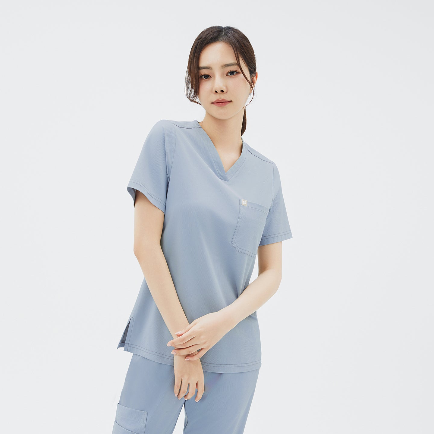Image of A close‑up, front three‑quarter portrait of the model with her hands gently crossed in front, focusing on the Women’s Single Pocket Scrub Top’s precise stitching around the V‑neck and its single front patch pocket.