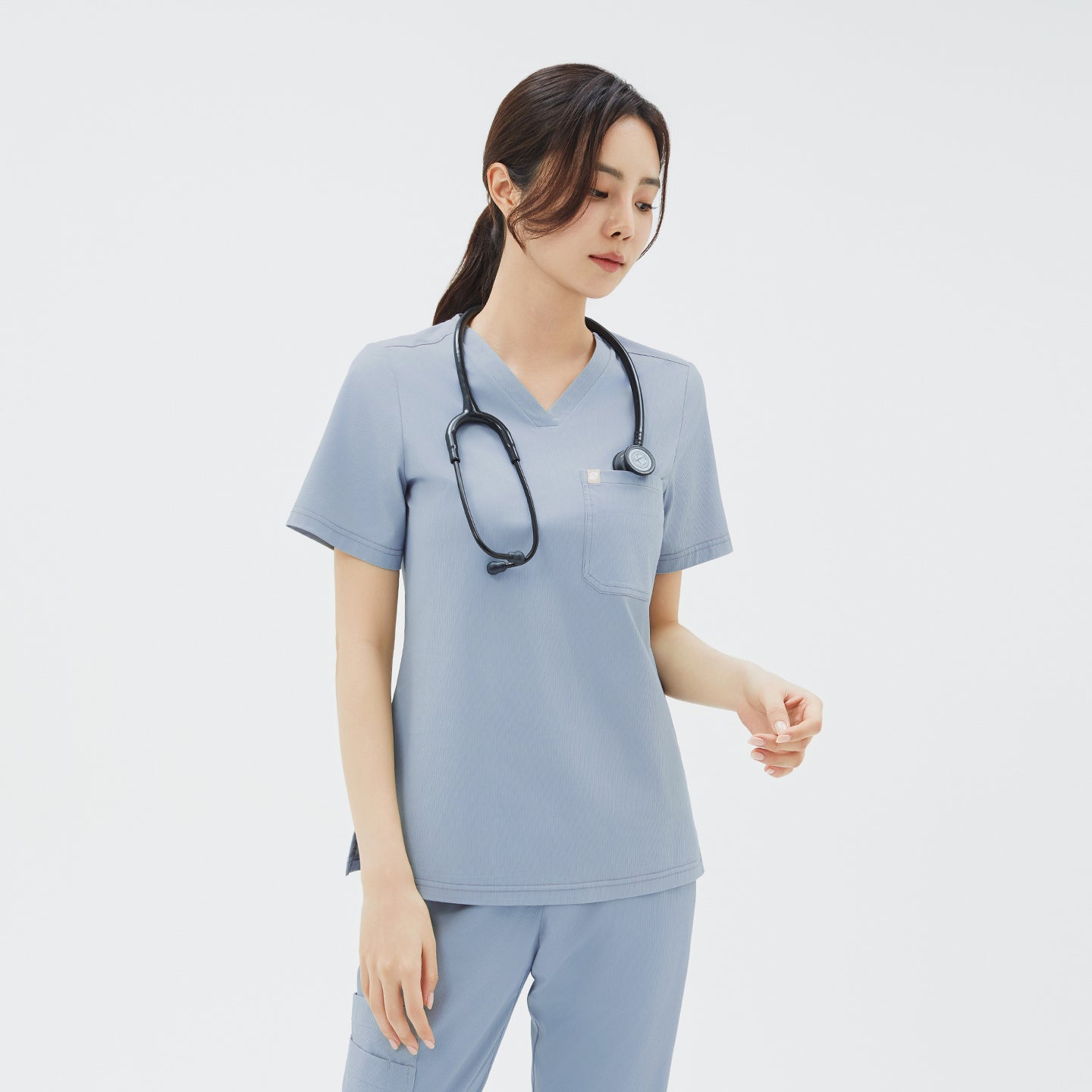A three‑quarter side view of the model gazing downward as she holds her stethoscope, showcasing the scrub top’s side slits and neat V‑neck detailing.