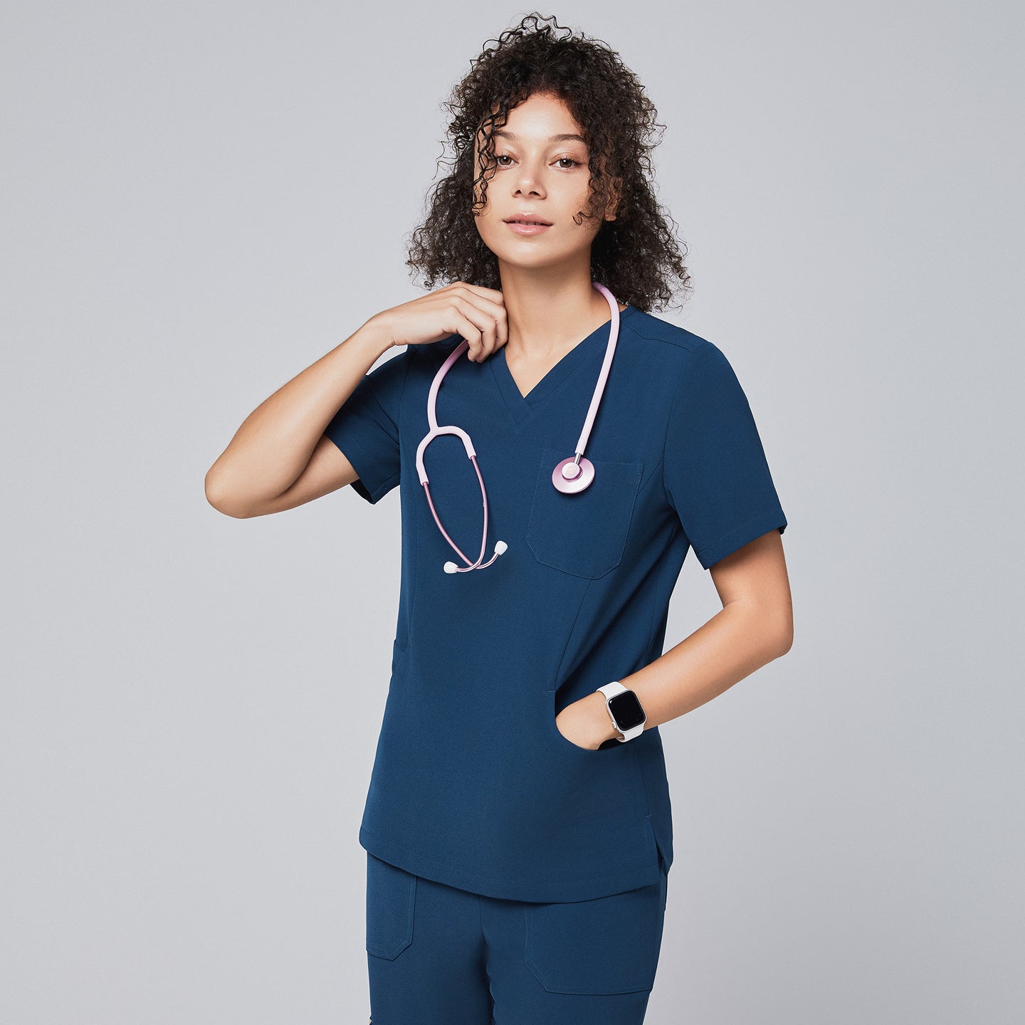 Woman wearing a dark blue V-neck scrub top with a pink stethoscope draped around her neck, posing confidently with one hand touching the stethoscope, against a light gray background,Dark Blue