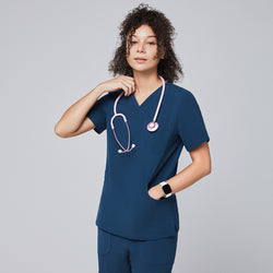 Image of Woman wearing a dark blue V-neck scrub top with a pink stethoscope draped around her neck, posing confidently with one hand touching the stethoscope, against a light gray background,Dark Blue